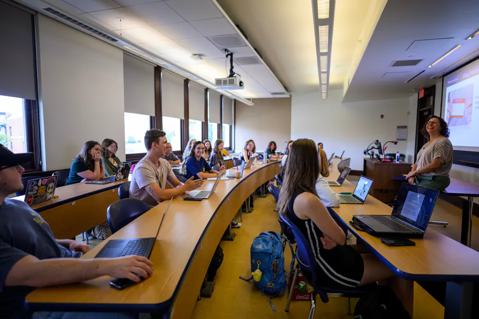A female instructor lectures to a class of college students seated at curved desks with laptops.