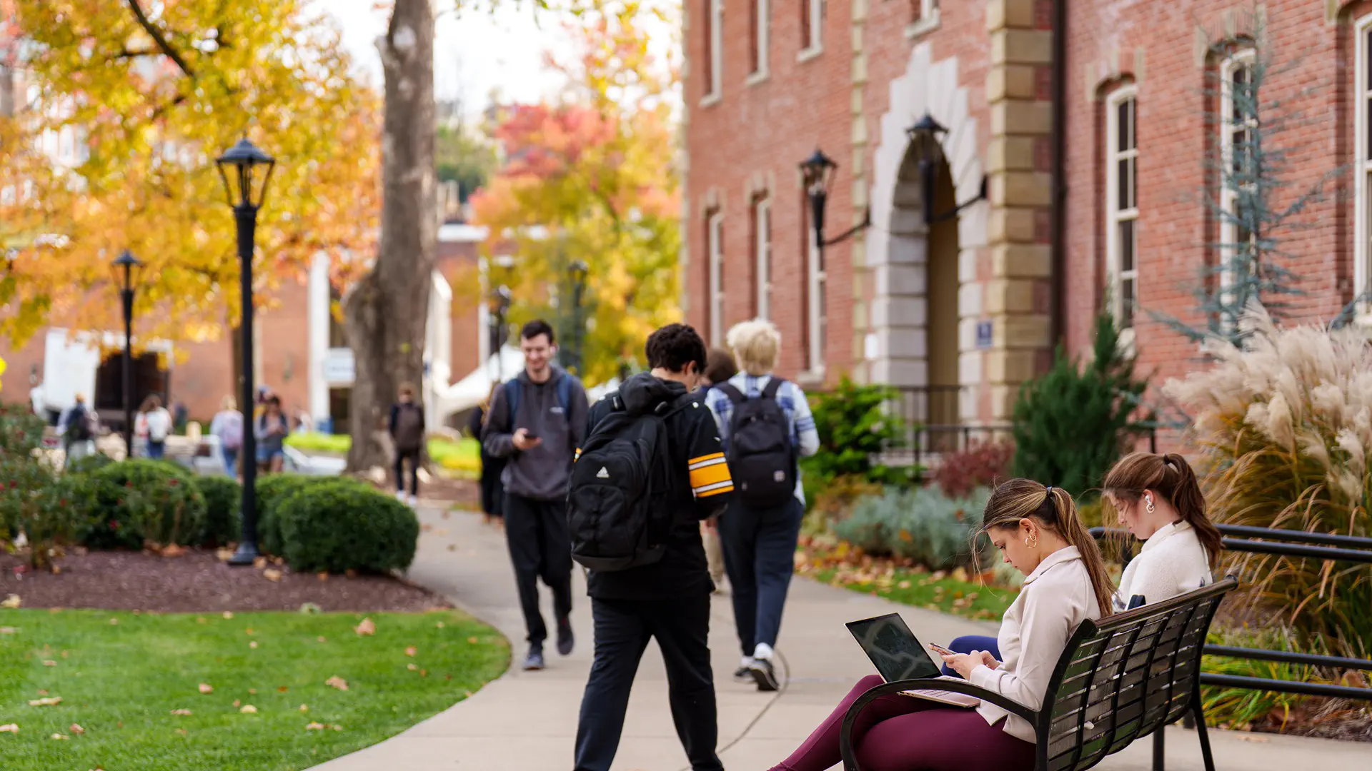 Two girls sitting on a bench in front of Martin Hall while other students walk by during Mountaineer Week on November 5, 2025, at West Virginia University in Morgantown, WV.