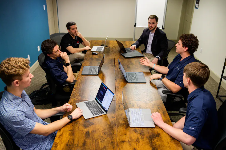 A group of men are engaged in a meeting around a large wooden table, each with a laptop in front of them.