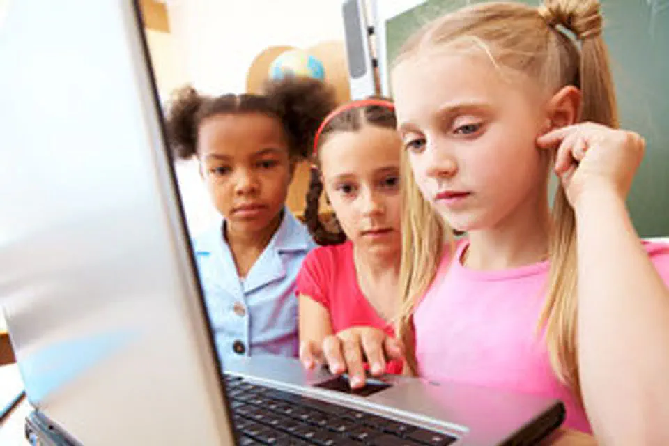 Three young girls are intently focused on a laptop screen in a classroom setting, suggesting a collaborative learning or discovery moment.